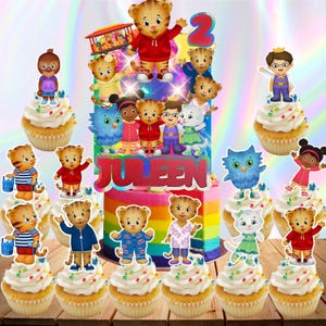 May include: A set of Daniel Tiger's Neighborhood themed cupcake toppers and a large cake topper featuring the characters, a trolley, and the name JULEEN in red letters. The toppers are displayed on cupcakes and a rainbow-colored cake.