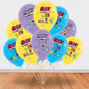 May include: A cluster of colorful balloons featuring the text "Abby Hatcher" and cartoon characters. The balloons are in shades of yellow, purple, and blue. They are tied to wooden sticks and arranged against a white wall and a wooden floor.