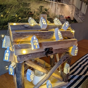 Könnte beinhalten: Eine rustikale dekorative Holzkiste, verziert mit einer Lichterkette aus warmweißen Feenlichtern und kleinen, weißen hausförmigen Lichtern. Die Kiste ist aus verwittertem Holz gefertigt und hat einen Metallverschluss. Ein weißer Kerzenhalter ist sichtbar.