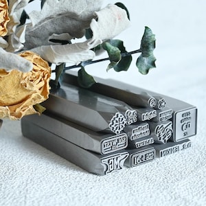 May include: A collection of rectangular metal stamps featuring various engraved designs and text including "SOL WEST 925", "los olivos metalworks", "ARGENT", "16 925", "MG", and "DR JENKIN". The stamps are arranged in a stack with a dried yellow rose and leaves.