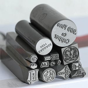 Puede incluir: Una colección de sellos metálicos de color gris oscuro con varios diseños y texto. Los sellos incluyen formas cilíndricas y rectangulares, con textos como "Cobber Co" y "DEBL BVCCVCE". Algunos sellos presentan diseños circulares y cuadrados.