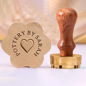 May include: A wooden pottery stamp with a flower-shaped design and the text "POTTERY BY SARAH" surrounding a heart. A separate stamp with a wooden handle and a brass base is also visible.