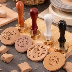 May include: A collection of wooden handled stamps with brass bases and various designs. The stamps are arranged with several clay impressions, some with text like "PLUM Boutique" and "Created by Claire". The handles are brown, red, white, and black.