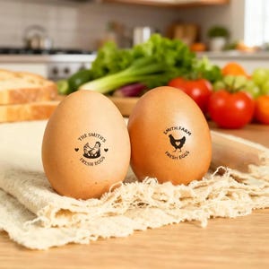 May include: Two brown eggs with stamped logos. One egg reads "The Smith's Fresh Eggs" with a hen illustration. The other egg reads "Smith Farm Fresh Eggs" with a chicken illustration. The eggs are on a white cloth, with vegetables in the background.