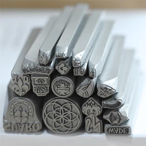 Puede incluir: Un conjunto de sellos de metal plateado con varios diseños, incluyendo una flor de lis, un trébol de cuatro hojas y patrones geométricos. Los sellos están dispuestos en una pila ordenada, mostrando sus grabados detallados.