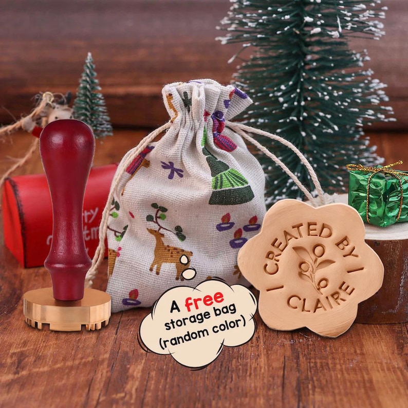 May include: A dark red wooden handle wax seal stamp with a brass base, a beige drawstring storage bag featuring a deer and tree print, and a flower-shaped wax seal impression with the text "CREATED BY I CLAIRE" and a small leaf design.