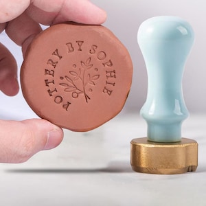 Könnte beinhalten: Ein Terrakotta-Töpferstempel mit dem Text "POTTERY BY SOPHIE", der ein Blumendesign umrahmt. Ein hellblauer und goldener Stempel befindet sich neben dem Töpferstempel.