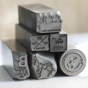 May include: A collection of six silver-toned metal stamping tools with various engraved designs and text. The stamps feature images of a donkey, the letters B and M, and text including Echo Lake Mint, Butler Mint Fine Copper 999, and Cu Copper.