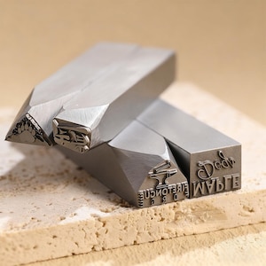 Op de afbeelding: Close-up van verschillende metalen stempels met diverse ontwerpen en tekst. Eén stempel heeft een pootafdruk, een andere de letters "HE", en andere tonen de woorden "Dad" en "Wife". De stempels liggen op een getextureerd oppervlak.