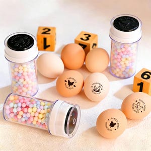 May include: An assortment of brown and white eggs, some stamped with "Fresh Eggs" and a chicken logo, alongside clear plastic containers filled with colorful spherical candies. Wooden number blocks (1, 2, 3, 6) are also present.
