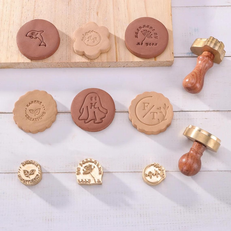 May include: A collection of wax seal stamps and stamped clay discs featuring various designs including a dolphin, a ghost, floral motifs, and text such as 'Karens Creations' and 'Handmade by Me'. The set includes two wooden handled brass stamps.