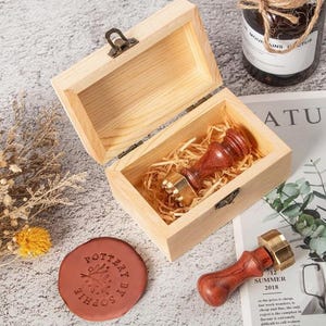 May include: A wooden box containing a wax seal stamp with a dark wood handle and brass head. A second matching stamp lies outside the box next to a round, reddish-brown wax seal featuring the embossed text "POTTERY BY SOPHIE" and a leaf design.