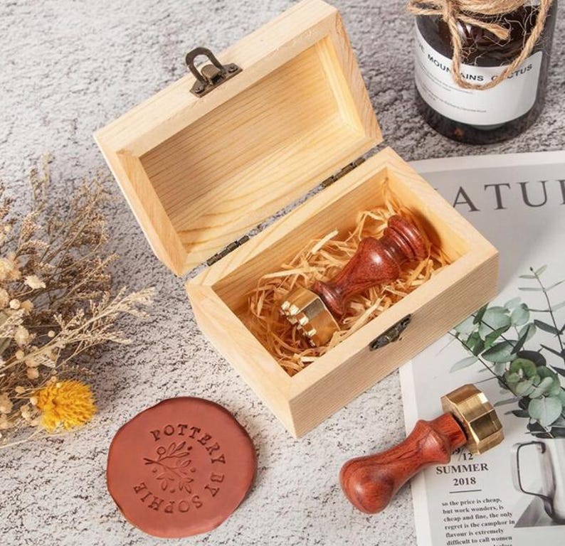 May include: A wooden box containing a wax seal stamp with a dark wood handle and brass head. A second matching stamp lies outside the box next to a round, reddish-brown wax seal featuring the embossed text "POTTERY BY SOPHIE" and a leaf design.