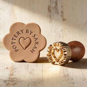 Puede incluir: Un sello de cerámica en forma de flor, de color marrón claro, con el texto "POTTERY BY SARAH" y un diseño de corazón. También hay un sello de metal dorado con un diseño de corazón y un mango de madera.