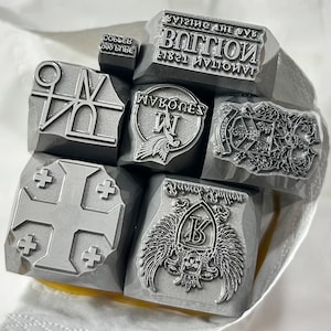 Puede incluir: Un conjunto de sellos de metal gris con varios diseños y texto. Los sellos presentan símbolos, cruces y textos como "VISITING THE BOTTOM" y "WYKONES". Los sellos están dispuestos en un paño blanco.