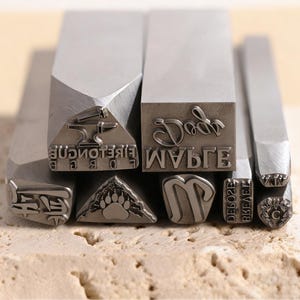 Könnte beinhalten: Eine Sammlung von Metallstempeln mit verschiedenen Designs und Texten. Die Stempel zeigen einen Amboss, einen Pfotenabdruck, das Wort "Dad", den Buchstaben "M" und andere Symbole und Buchstaben. Die Stempel sind silberfarben und haben eine rechteckige Form.