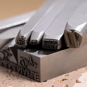 May include: Close-up of several metal letter stamps with various fonts and designs. The stamps are silver-coloured and have different letter and symbol impressions on the bottom. Some stamps have floral designs. The stamps are arranged on a metal block.