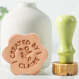 May include: A light green wax seal stamp with a brass base and a light green handle. A flower-shaped stamp with the text "I CREATED BY I CLAIRE" is next to a white vase.