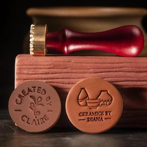 Puede incluir: Un sello de cerámica con mango de madera y cabeza de latón, apoyado sobre un bloque de terracota. También se ven dos sellos de arcilla de terracota, uno con el texto "Created by Claire" y otro con el texto "Ceramics by Diana".