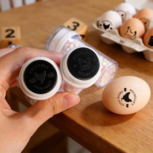 May include: Two egg stamps with black ink and a chicken design are held in front of a wooden surface. The stamps are next to an egg with a chicken design and the words "MICHELLE'S FRESH EGGS". A carton of eggs with chicken designs is in the background.