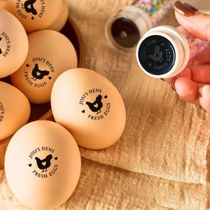 May include: A collection of beige eggs, each bearing a black hen logo and the text "Josi's Hens Fresh Eggs." A matching stamp is held in the frame. The eggs are presented in a wooden container, evoking a rustic, farm-fresh aesthetic.