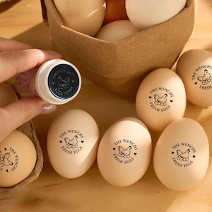 May include: A collection of fresh eggs, some stamped with "THE WENCHS FRESH EGGS" in black ink. A stamp with the same design is held nearby. The eggs are in a brown basket and on a wooden surface.