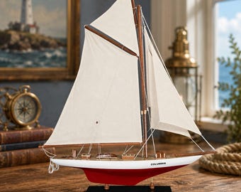 May include: A model sailing boat with a white hull, a red bottom, and off-white fabric sails. The wooden deck features small details, and the name COLUMBIA is printed in black on the side of the hull. The boat is mounted on a black rectangular base.