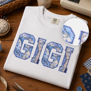 May include: A white t-shirt featuring the word GIGI in large block letters. The letters are filled with a blue and white floral and checkered patchwork pattern. A circular inset shows a close-up of the fabric texture on the letter G.