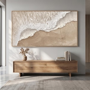May include: A large rectangular framed artwork depicting a beach scene with textured white waves and tan sand. The artwork is mounted on a light gray wall above a wooden cabinet.