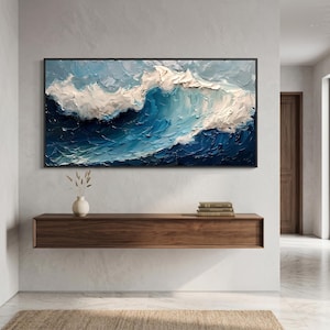 Puede incluir: Gran pintura abstracta enmarcada de una ola en tonos azules y blancos. La obra presenta gruesas pinceladas, creando un efecto texturizado. La pintura se exhibe sobre una consola de madera en un entorno interior moderno.