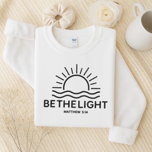 May include: White sweatshirt with a black graphic of a sun over waves and the text "BE THE LIGHT" and "MATTHEW 5:14". The sweatshirt is folded on a cream-colored surface.