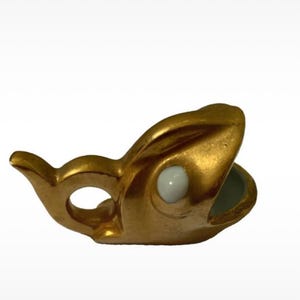 Może przedstawiać: Mała, złotego koloru ceramiczna figurka w kształcie stylizowanej ryby z otwartym pyskiem. Ryba ma gładkie, metaliczne złote wykończenie oraz pojedynczy, wyraźny biały okrągły detal oka z boku, z widocznym pustym wnętrzem.