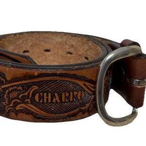 May include: A brown leather belt with a silver buckle. The belt features embossed floral designs and the word "CHARRO". The belt is rolled up, showcasing the intricate details and craftsmanship.