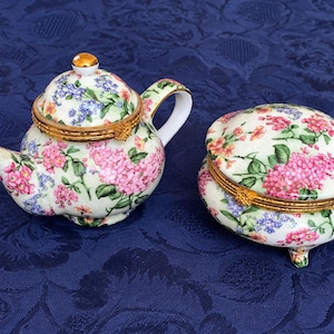 Può includere: Una teiera in porcellana e un portagioie rotondo abbinato, entrambi con base color crema e un vivace motivo floreale di fiori rosa, blu e verdi. Ogni pezzo include un coperchio incernierato con bordo e chiusura in metallo color oro.