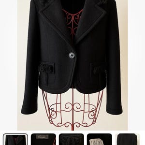Può includere: Giacca nera in lana con chiusura a un bottone, colletto a revers e frange decorative sulle spalle e sulle tasche. La giacca è esposta su una gruccia di metallo rossa. Il testo sull'immagine dice "Eureka".