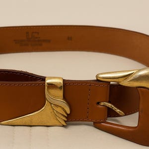 以下が含まれることがあります： 金色のバックルが付いた茶色の革ベルト。バックルは、湾曲した形状と装飾的な要素を備えたユニークなデザインです。ベルトにはサイズ41と記載されています。