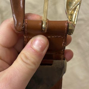 以下が含まれることがあります： 金色のバックルが付いた茶色の革ベルト。バックルは装飾的なデザインです。ベルトは手で持たれています。革には使用感が見られます。