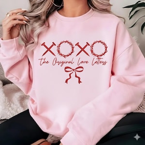 May include: Light pink sweatshirt with red graphic text and design. The design includes the letters "XOXO" with nails and a crown of thorns, and the words "The Original Love Letters" with a bow.