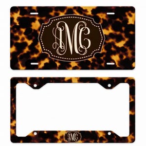 May include: A license plate and license plate frame with a tortoiseshell pattern. The license plate has a brown oval with the white letters "MC". The frame has the same pattern and a small oval with the letters "MC".