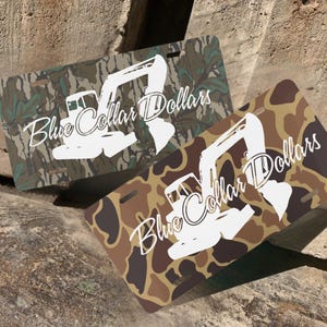 May include: Two license plates with a camouflage pattern. One plate has a green and brown camouflage pattern, and the other has a brown and tan camouflage pattern. Both plates have a white excavator graphic and the words "Blue Collar Dollars".