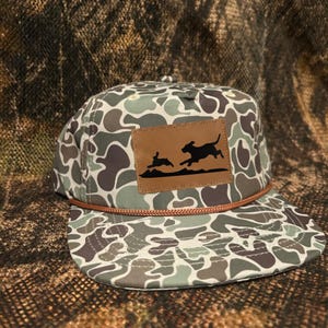 May include: Camouflage pattern baseball cap with a brown leather patch featuring a black silhouette of a dog chasing a rabbit. The cap has a brown rope detail along the brim and a flat bill.