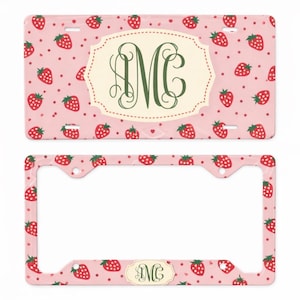 May include: Pink license plate and frame set with a strawberry pattern. The license plate features a large, ornate initial "M" in green within a cream-colored oval. The frame has a matching strawberry design.