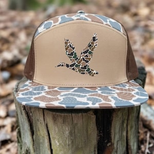 May include: A brown and tan trucker hat with a camouflage pattern on the bill and top. The front panel is tan with a camouflage duck graphic. The hat has a brown mesh back.