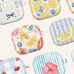 May include: A collection of colorful AirPod cases with various designs. Patterns include lemons, strawberries, hibiscus flowers, and blue stripes. Some cases feature bows and floral arrangements. The cases are white, blue, pink, and yellow.