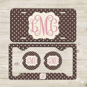 May include: A brown license plate and frame set with pink polka dots. The license plate features a white, scalloped design with a pink monogram. The set includes two matching car coasters and a license plate frame.