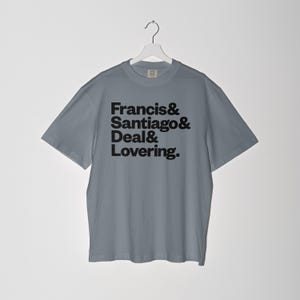May include: A gray t-shirt with the names "Francis & Santiago & Deal & Lovering" printed in black. The shirt is hanging on a white hanger against a white background. The t-shirt has short sleeves and a crew neck.