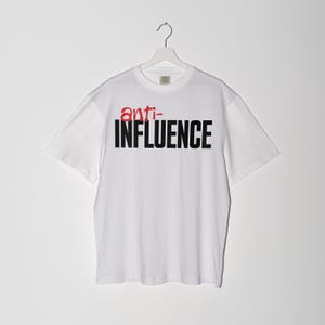 Peut inclure: T-shirt blanc avec l'inscription "anti-INFLUENCE" imprimée en noir et rouge sur le devant. Le t-shirt est suspendu à un cintre blanc sur fond blanc. Le t-shirt a des manches courtes.