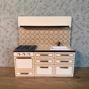 Op de afbeelding: Een miniatuurkeuken in wit en bruin. Het heeft een fornuis, oven, gootsteen en kasten. De achterwand heeft een patroon. De keuken is gemaakt van hout en is ontworpen voor een poppenhuis.