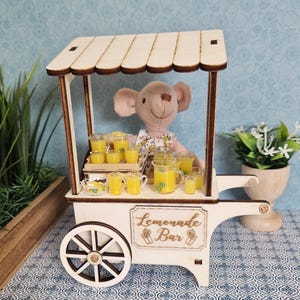Könnte beinhalten: Ein Miniatur-Holz-Limonadenstand mit beigem Baldachin und einem Schild mit der Aufschrift "Lemonade Bar". Der Stand zeigt eine Plüschmaus und mehrere Gläser Limonade. Der Wagen hat einen weißen Körper und ein Holzrad.
