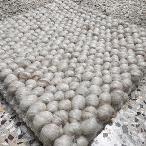 May include: A square, cream-coloured rug with a textured surface. The rug is made of tightly packed, rounded fibres, creating a bumpy, tactile appearance. The rug's neutral colour and unique texture make it a versatile home decor item.